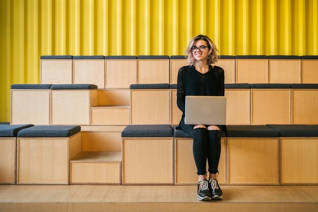 Jeune femme séduisante assise dans une salle de conférence, travaillant sur un ordinateur portable, portant des lunettes, auditorium moderne, l'éducation des étudiants en ligne, pigiste, souriant, démarrage chez les adolescentes, à la recherche à huis clos, heureux