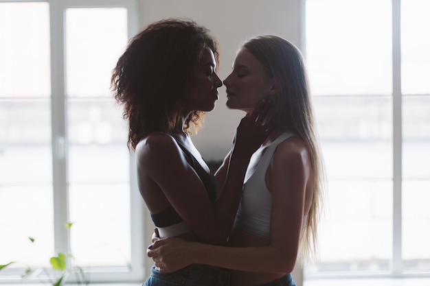 Photo gratuite jeune femme séduisante aux cheveux blonds et femme afro-américaine aux cheveux bouclés noirs se serrant rêveusement tout en passant du temps ensemble à la maison avec des fenêtres en arrière-plan