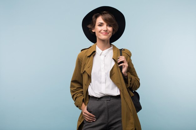 Jeune femme séduisante aux cheveux courts foncés en trench-coat et chapeau avec sac à dos regardant joyeusement à huis clos sur fond bleu isolé