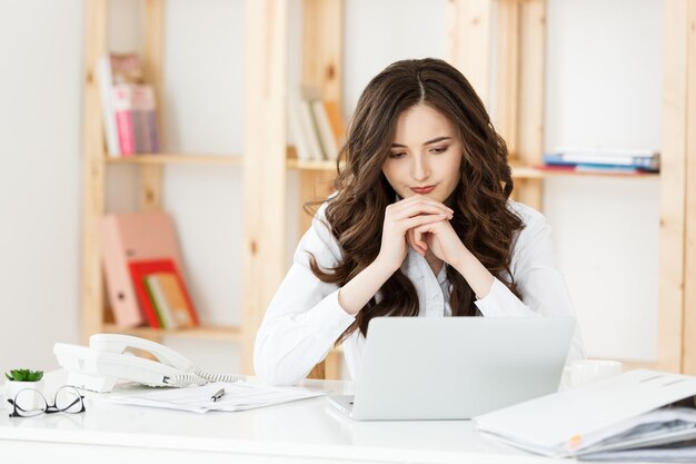 Jeune femme séduisante à un bureau moderne travaillant avec un ordinateur portable et penser à quelque chose