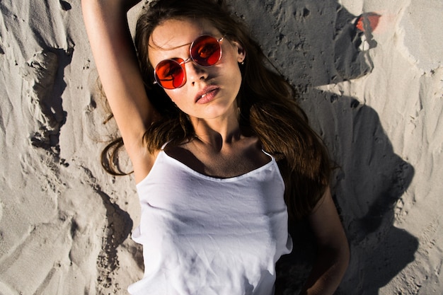 Une jeune femme séduisante dans des lunettes de soleil rouges repose sur du sable blanc