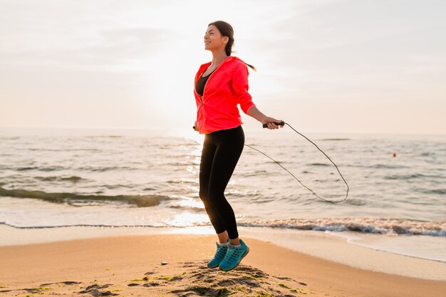 Jeune femme séduisante, faire des exercices de sport au lever du soleil du matin sur la plage de la mer en vêtements de sport, mode de vie sain, écouter de la musique sur les écouteurs, porter une veste coupe-vent rose, sauter en corde à sauter