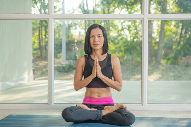 Jeune femme séduisante pratiquant le yoga, assise à Padmasana, exercice, pose de lotus, namaste, entraînement, portant des vêtements de sport, pantalon noir, intérieur pleine longueur, près de la fenêtre du sol.