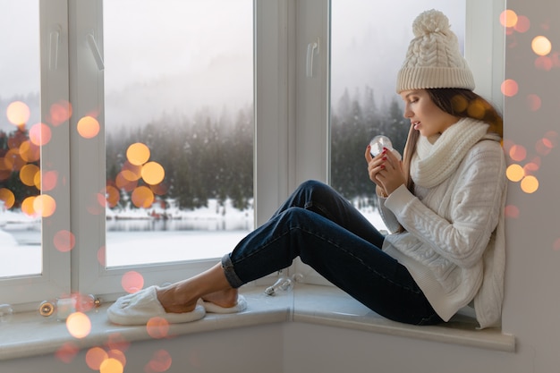 Jeune femme séduisante en pull tricoté blanc élégant, écharpe et chapeau assis à la maison sur le rebord de la fenêtre à Noël tenant boule de neige en verre décoration actuelle, vue sur la forêt d'hiver, lumières bokeh