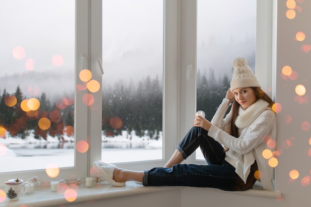 Jeune femme séduisante en pull tricoté blanc élégant, écharpe et chapeau assis à la maison sur le rebord de la fenêtre à Noël tenant boule de neige en verre décoration actuelle, vue sur la forêt d'hiver, lumières bokeh