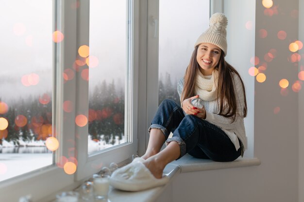 Jeune femme séduisante en pull tricoté blanc élégant, écharpe et chapeau assis à la maison sur le rebord de la fenêtre à Noël tenant boule de neige en verre décoration actuelle, vue sur la forêt d'hiver, lumières bokeh