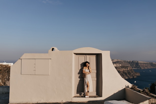 Jeune femme séduisante en robe midi pose près de la maison beige