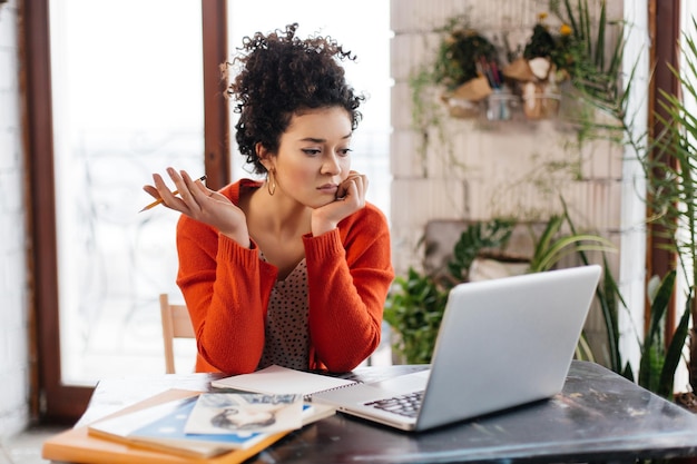 Photo gratuite jeune femme sérieuse aux cheveux bouclés noirs assis à la table appuyée sur la main tout en regardant avec étonnement sur un ordinateur portable dans un atelier moderne avec de grandes fenêtres en arrière-plan