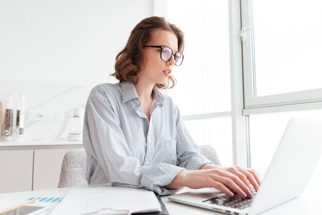 Jeune femme sérieuse en chemise rayée en tapant un e-mail à son patron tout en étant assis sur le lieu de travail dans un appartement lumineux