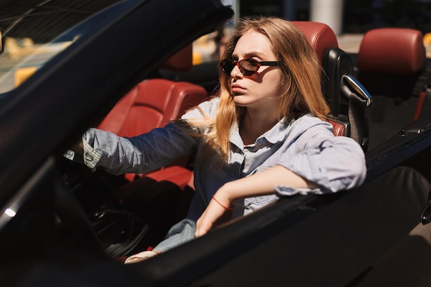 Jeune femme sérieuse à lunettes de soleil conduisant pensivement une voiture cabriolet en plein air