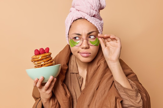 Photo gratuite une jeune femme sérieuse regarde attentivement de côté garde la main sur le bord des lunettes pose avec de délicieuses crêpes applique des taches vertes sous les yeux recouverts d'une couverture isolée sur fond marron