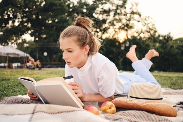 Jeune femme sérieuse séduisante allongée sur un plaid lisant attentivement un livre sur un pique-nique dans le magnifique parc de la ville