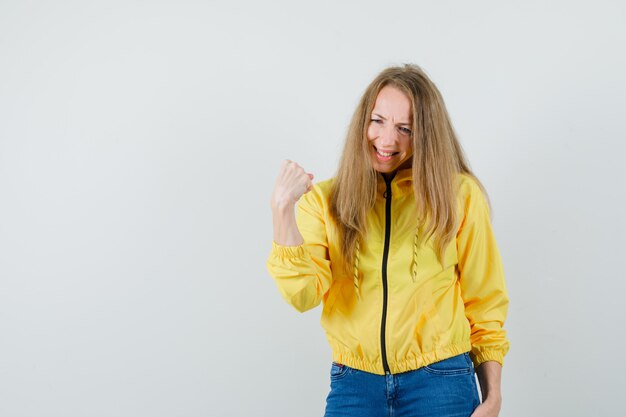 Jeune femme serrant le poing en blouson aviateur jaune et jean bleu et à la vue épuisée, de face.