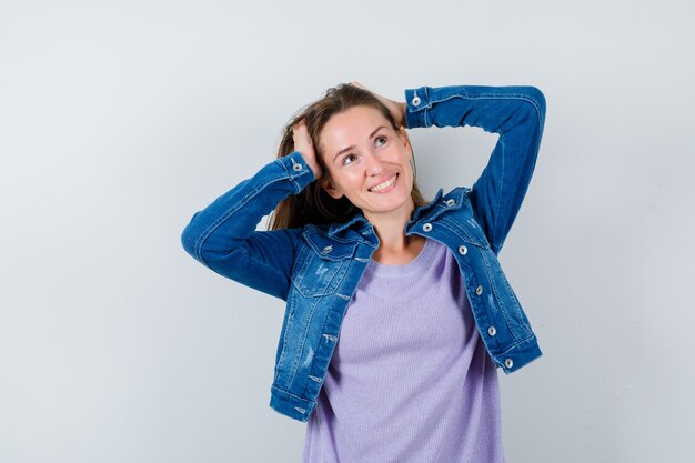 Jeune femme serrant la tête avec les mains, levant les yeux en t-shirt, veste et semblant heureuse. vue de face.