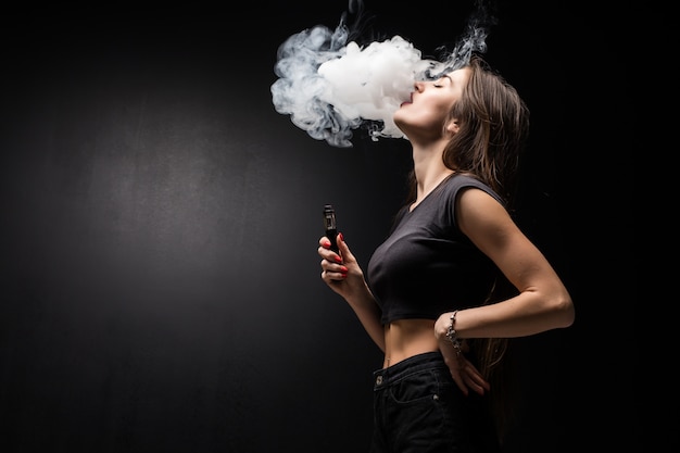 Jeune femme sexy vapote en fumant une cigarette électronique. nuage de vapeur sur le mur noir