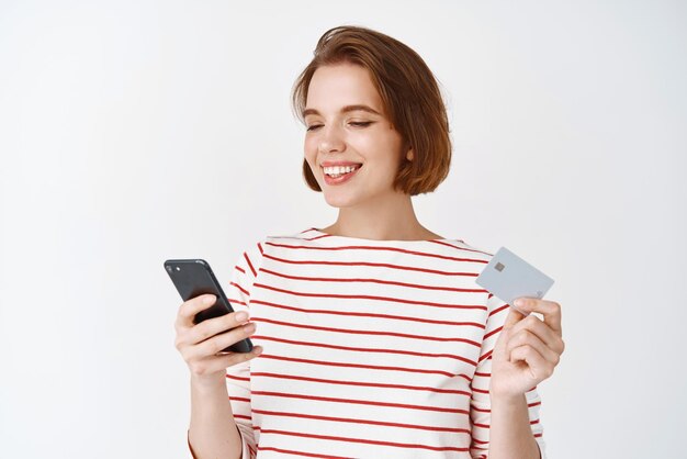 Jeune femme shopping en ligne avec carte de crédit en plastique et smartphone fille lisant l'écran du téléphone portable et souriant payant sur fond blanc internet