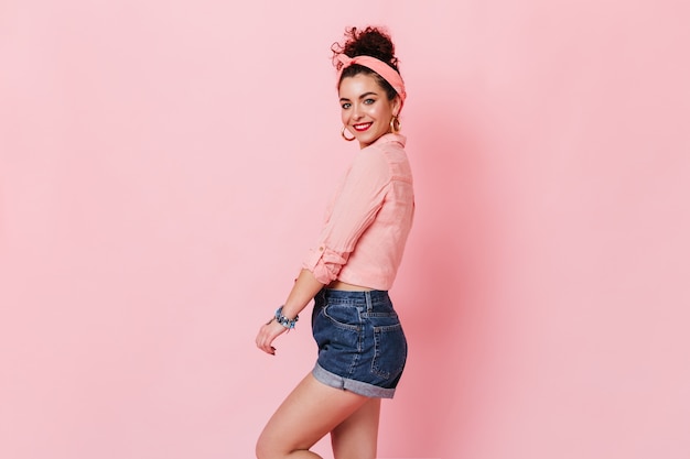 Jeune femme en short en jean, chemisier rose et bandeau mignon souriant sur un espace isolé.