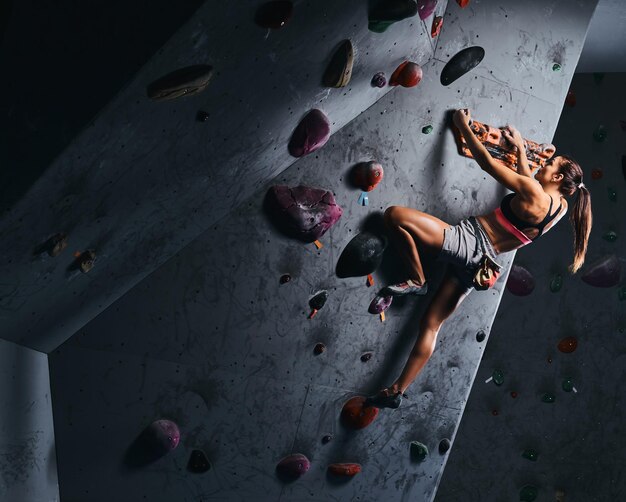 Jeune femme en short et soutien-gorge de sport exerçant sur un mur de bloc à l'intérieur.