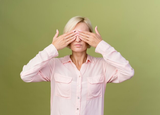 Jeune femme slave blonde couverte de yeux avec les mains isolées sur le mur vert olive