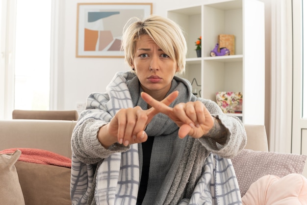 Jeune femme slave malade mécontente avec une écharpe autour du cou enveloppée dans un plaid croisant ses doigts ne faisant aucun signe assis sur un canapé dans le salon