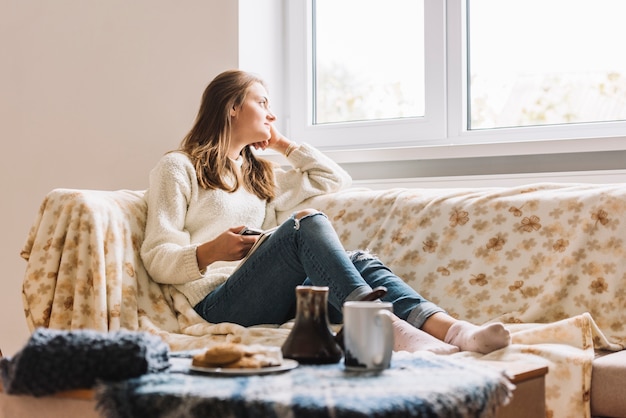 Jeune femme avec un smartphone sur un canapé près de table avec boisson et biscuits