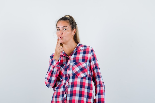 Jeune femme soufflant sur le pistolet à doigt en chemise à carreaux et à la confiance. vue de face.