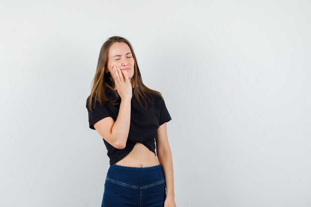 Jeune femme souffrant de maux de dents en chemisier noir, pantalon et à l'air troublé.