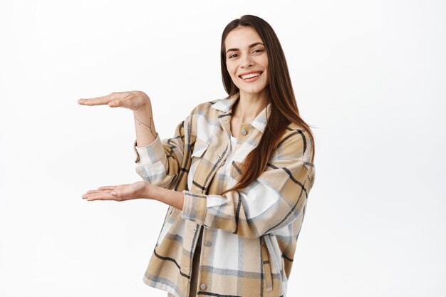 Une jeune femme souriante affiche un article dans ses mains, montrant le geste de la boîte, démontre le logo ou l'offre d'achat, mur blanc.