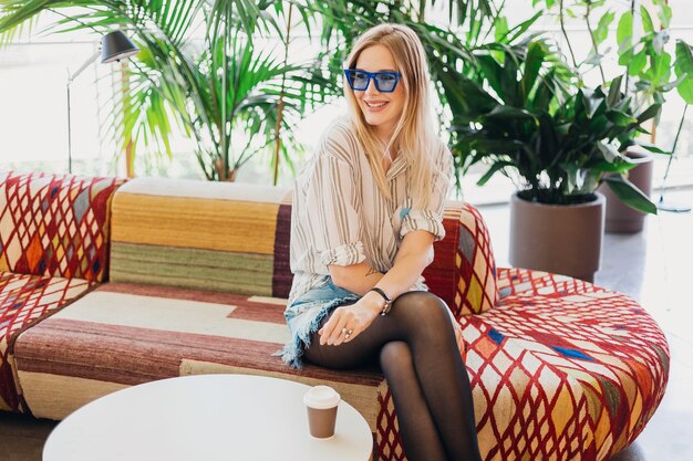 Jeune femme souriante assez élégante assise dans un café hipster sur un canapé portant une chemise et des lunettes de soleil bleues