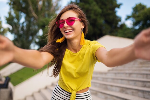 Jeune femme souriante assez élégante faisant selfie dans le parc de la ville, positive, émotionnelle, portant haut jaune, lunettes de soleil roses, tendance de la mode estivale, cheveux longs, s'amuser