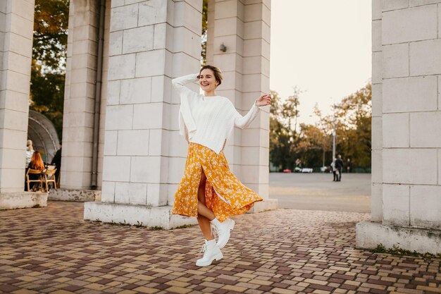 Jeune femme souriante assez heureuse en robe imprimée jaune et pull blanc tricoté