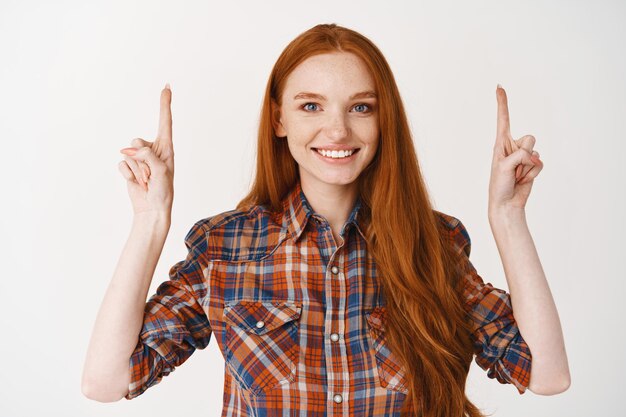 Jeune femme souriante aux longs cheveux roux et aux yeux bleus, pointant les doigts vers le haut avec un visage heureux
