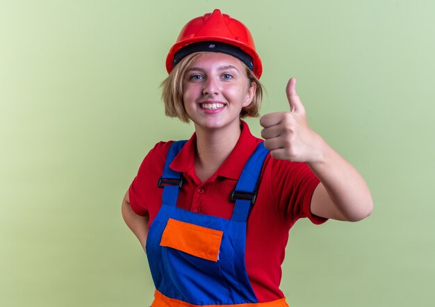 Jeune femme souriante de constructeur en uniforme montrant le pouce vers le haut isolé sur un mur vert olive