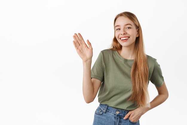 Jeune femme souriante et disant bonjour, vous saluant, bienvenue aux gens, debout dans des vêtements d'été contre un mur blanc