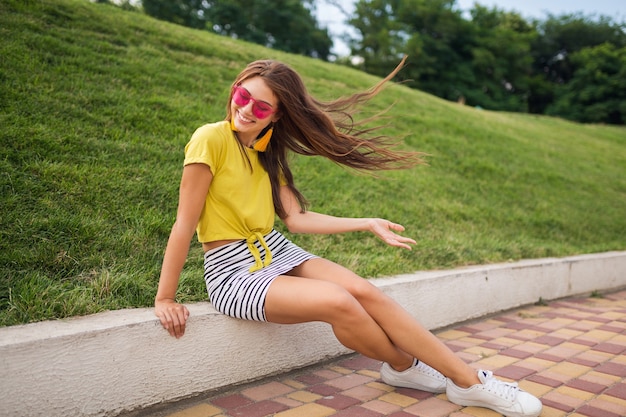 Photo gratuite jeune femme souriante élégante et attrayante s'amusant dans le parc de la ville, positive, émotionnelle, portant haut jaune, mini jupe rayée, lunettes de soleil roses, baskets blanches, tendance de la mode de style d'été, longues jambes