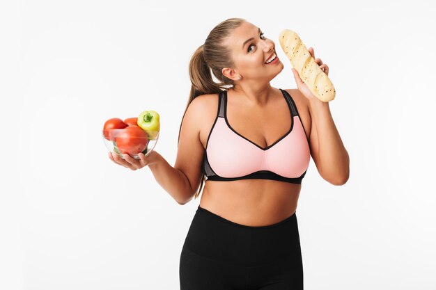 Jeune femme souriante avec un excès de poids dans un haut sportif tenant un bol avec des légumes mais en choisissant du pain tout en regardant rêveusement à huis clos sur fond blanc