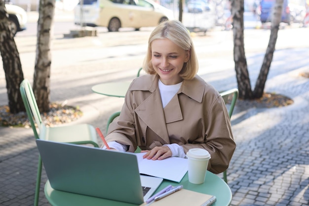 Photo gratuite une jeune femme souriante et heureuse dans une tranchée assise dans un café de rue en plein air, buvant du café en freelance.