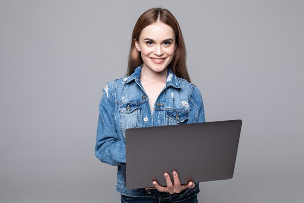 Jeune femme souriante heureuse dans des vêtements décontractés tenant un ordinateur portable et envoyer un e-mail à son meilleur ami isolé sur mur gris