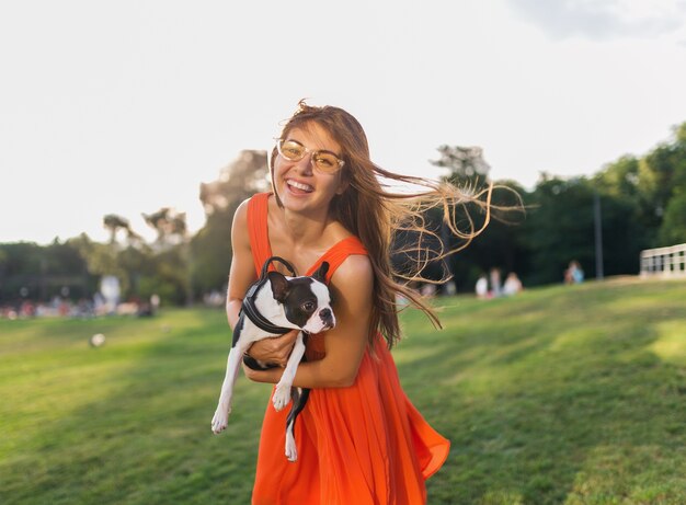 Jeune femme souriante heureuse en robe orange s'amusant à jouer avec un chien dans le parc, style d'été, bonne humeur