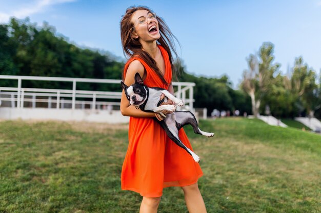 Jeune femme souriante heureuse en robe orange s'amusant à jouer avec un chien dans le parc, style d'été, bonne humeur