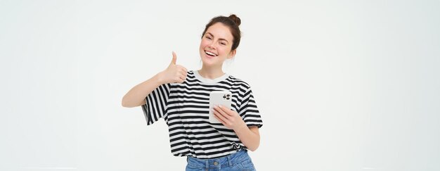 Photo gratuite une jeune femme souriante montrant le pouce levé en signe d'approbation recommande que l'application tienne le smartphone semble satisfaite