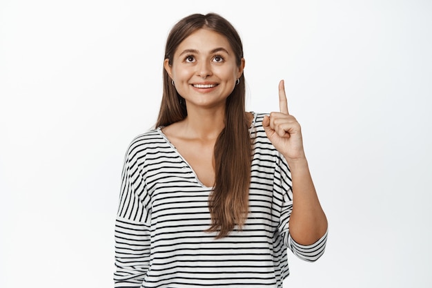 Jeune femme souriante pointant vers le haut avec une expression de visage heureux sur blanc.