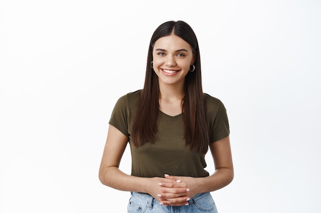 Jeune femme souriante prête à offrir son service, comment puis-je vous aider, les mains serrées et l'air amical à l'avant, mur blanc