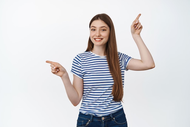 Une jeune femme souriante suggère deux options, pointant les doigts vers le haut et vers la gauche, montrant des variantes, debout sur fond blanc