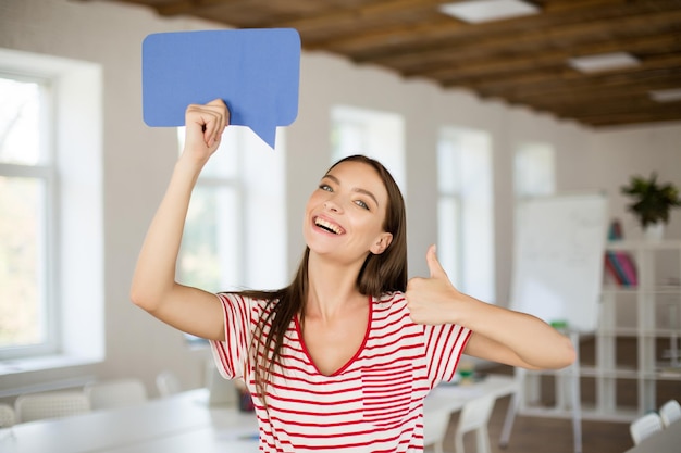 Jeune femme souriante en T-shirt rayé regardant joyeusement à huis clos tenant l'icône de message papier dans la main près de la tête tout en montrant le geste du pouce vers le haut au bureau