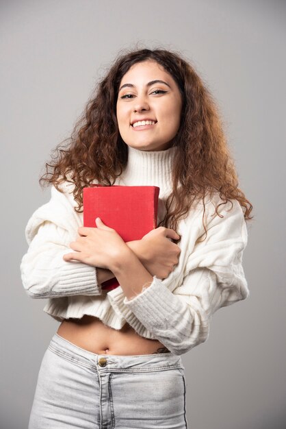 Jeune femme souriante tenant un livre rouge sur un mur gris. Photo de haute qualité