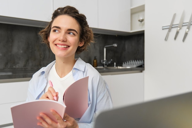 Une jeune femme souriante travaille à domicile écrit dans son journal et prend des notes dans le planificateur de journal se trouve à proximité