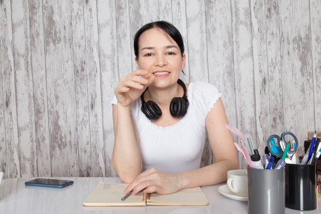 jeune femme, sourire, dans, chemise blanche, café buvant, écouter musique, dans, noir, écouteurs, écrire, notes, sur, mur gris