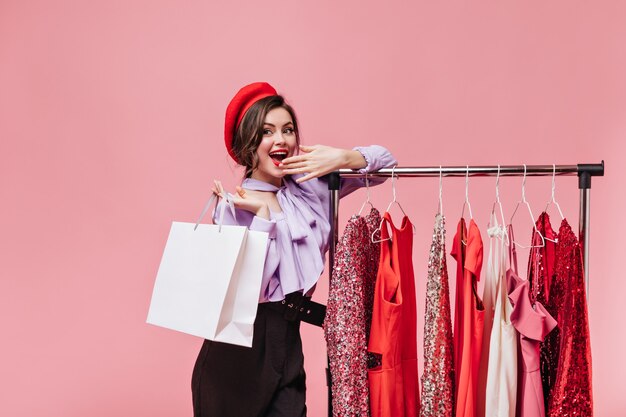 Jeune femme sourit et se couvre la bouche lors de ses achats. Dame en béret pose près de stand avec des robes de fantaisie.