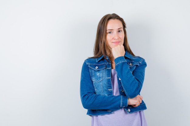 Jeune femme soutenant le menton sur le poing en t-shirt, veste en jean et semblant positive. vue de face.